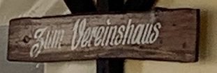 Schild mit der Aufschrift "Zum Vereinshaus" auf einem Holzbrett.